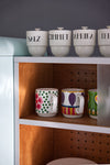 Set of canisters labeled with spices on a shelf above colorful mugs in a kitchen.